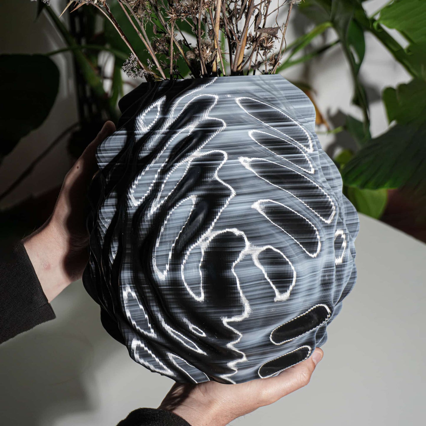 Person holding a unique vase with a black and white pattern against a blurred plant background