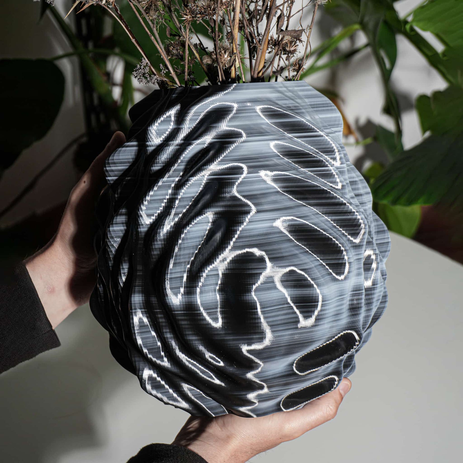 Person holding a unique vase with a black and white pattern against a blurred plant background
