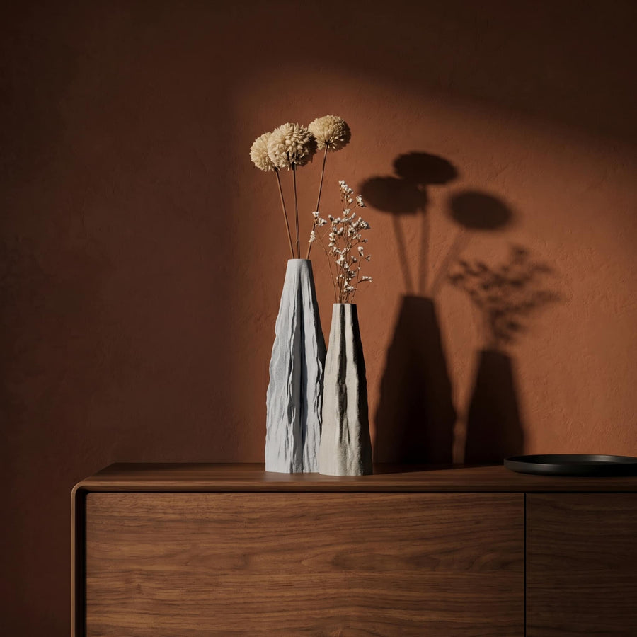 Peak Duo vases with dried flowers on a wooden surface against a warm brown wall.