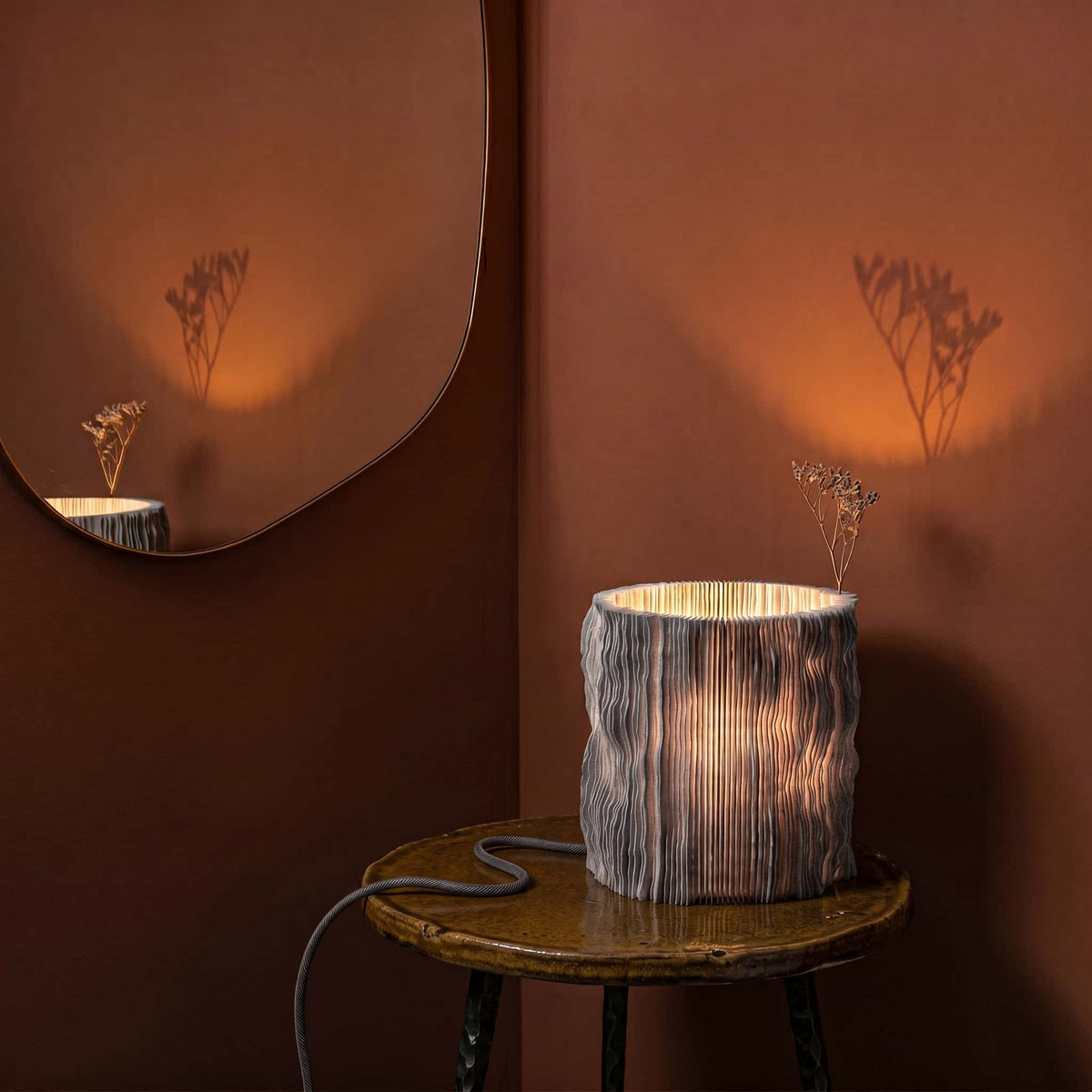 Profilum lamp on a small table with a mirror reflecting plants in the lamp folds against a warm wall.
